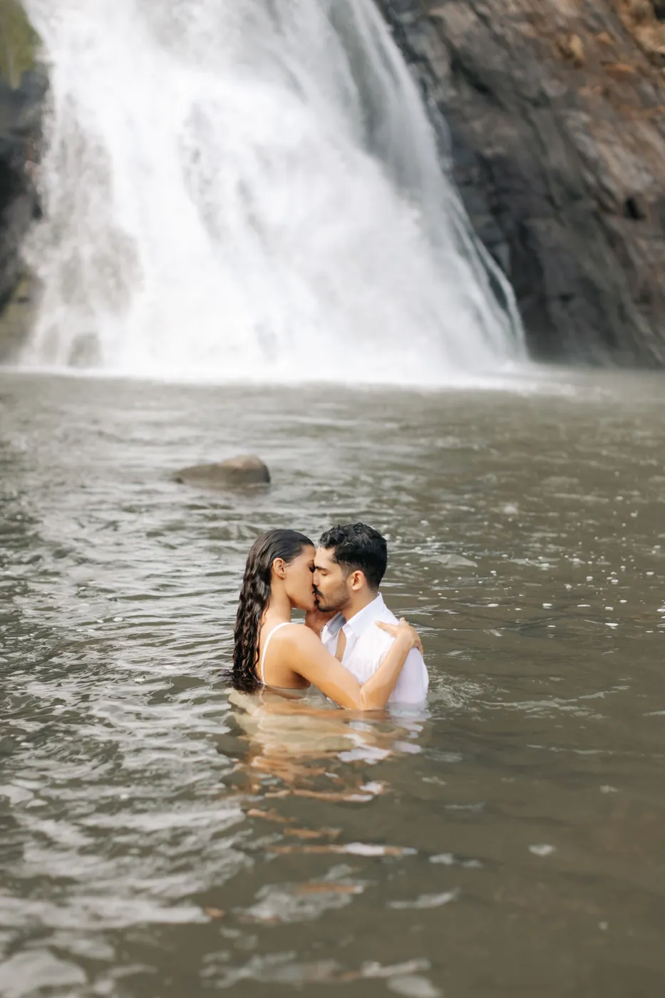 Casal se beijando dentro da água, com a cachoeira ao fundo