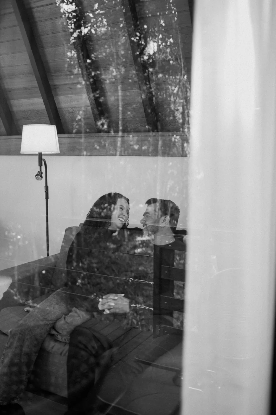 Casal rindo no sofá, visto pelo reflexo do vidro, em preto e branco